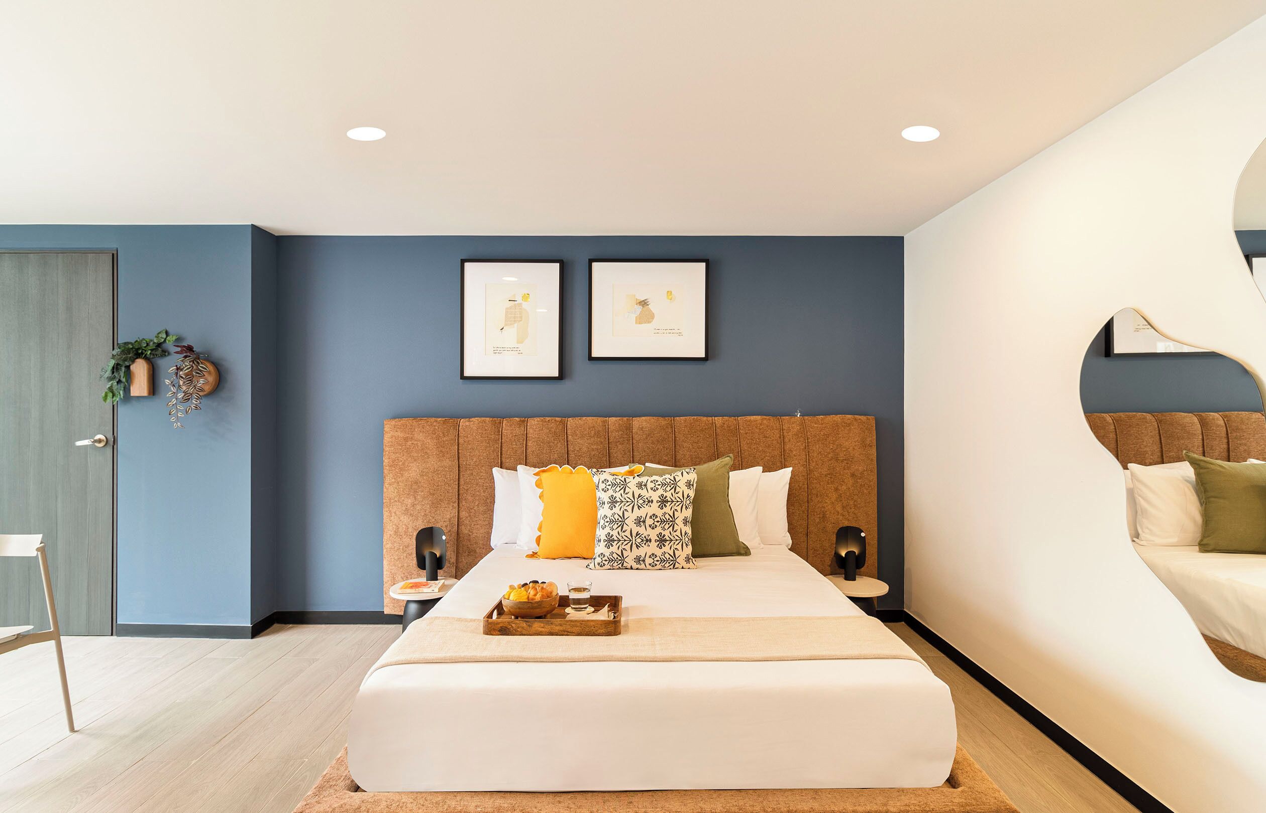 Een moderne slaapkamer met een groot bed, een hoofdeinde, twee ingelijste kunstwerken, een nachtkastje met een fruitschaal, en een deur met een plant.