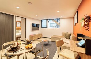 Una sala de estar moderna con comedor, un conjunto de sofás, un televisor montado en la pared y una escalera que lleva a un piso superior.