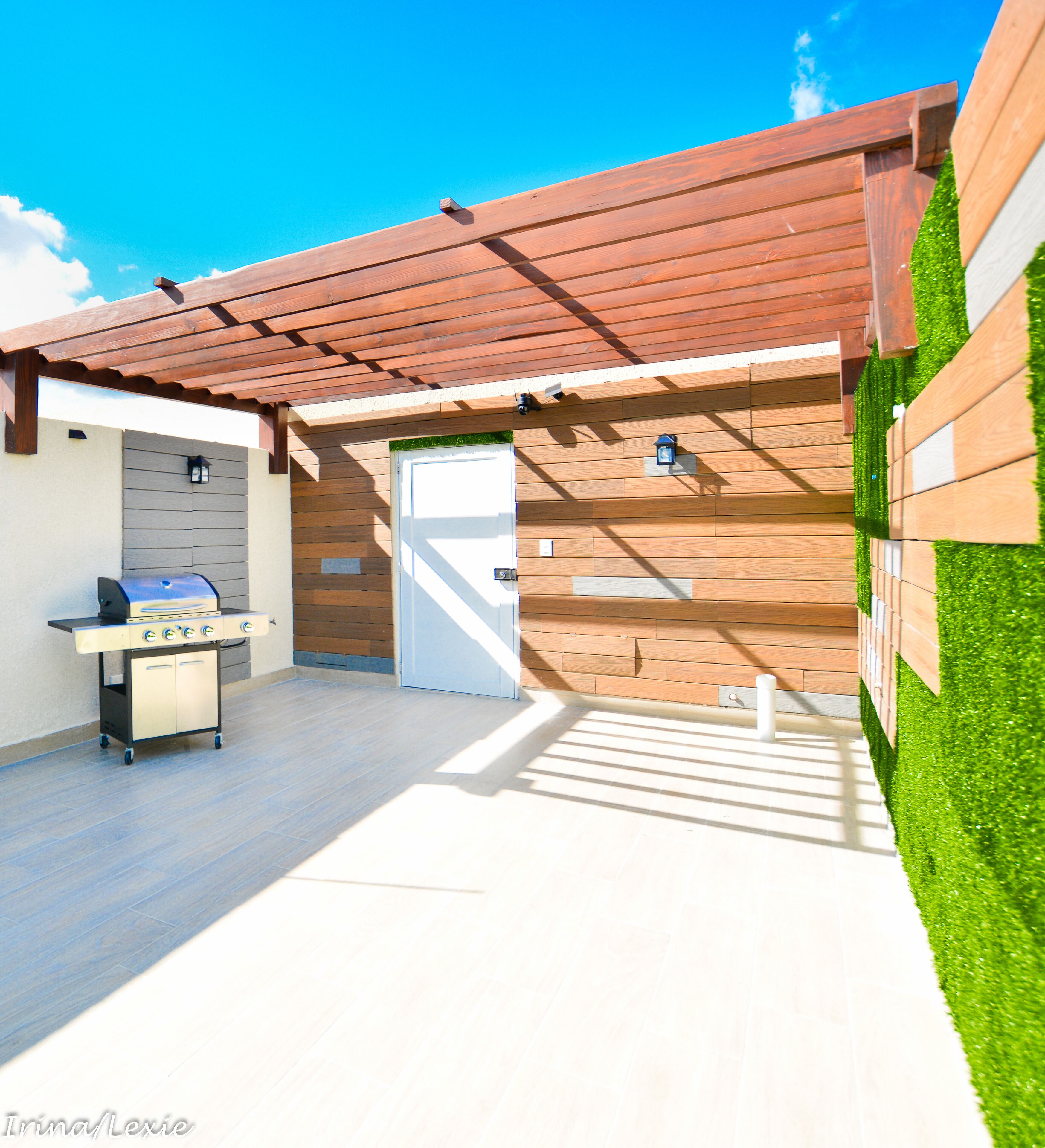 Een overdekt terras met een barbecue, een deur en een muur met houten panelen en kunstgras.