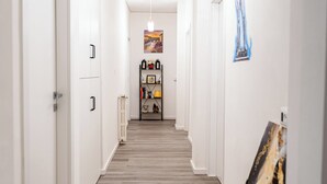 Un couloir avec un plancher en bois, des murs blancs et un luminaire au plafond. Il y a une porte sur la gauche et une étagère contenant divers objets sur la droite.