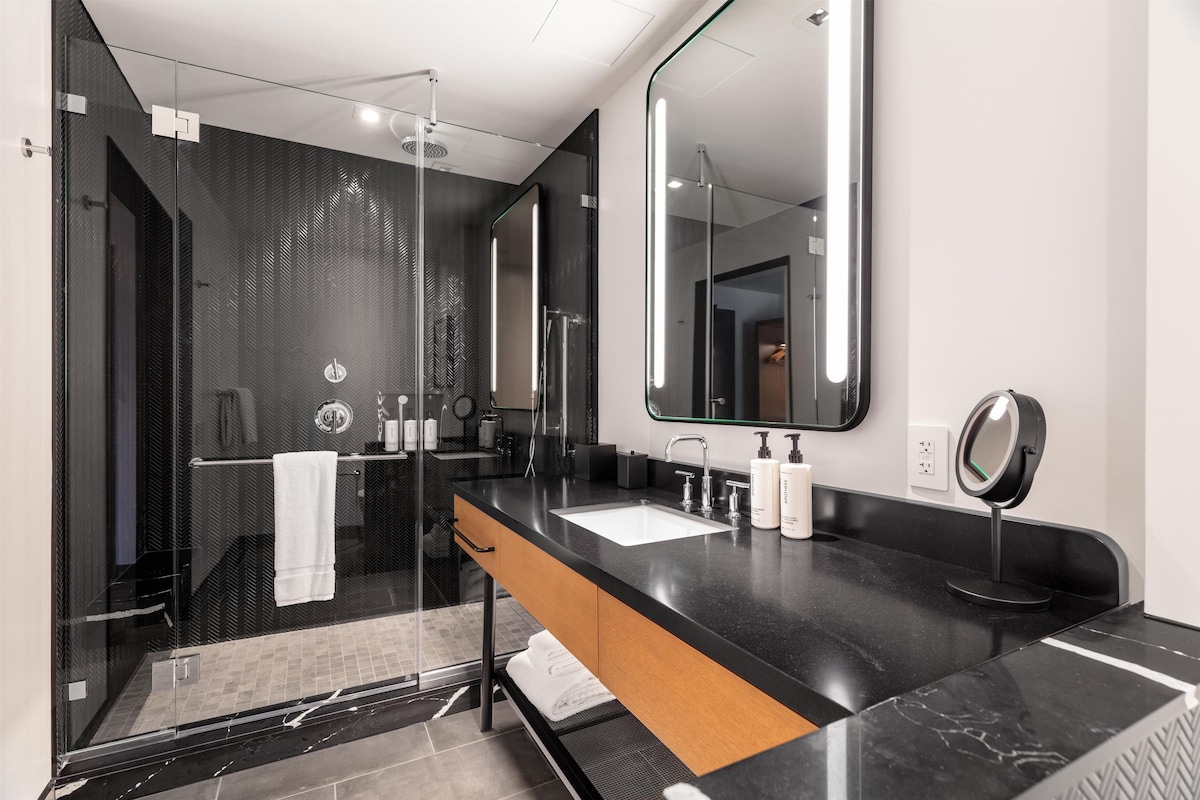 A modern bathroom with a black marble countertop, a large mirror, and a glass shower enclosure.