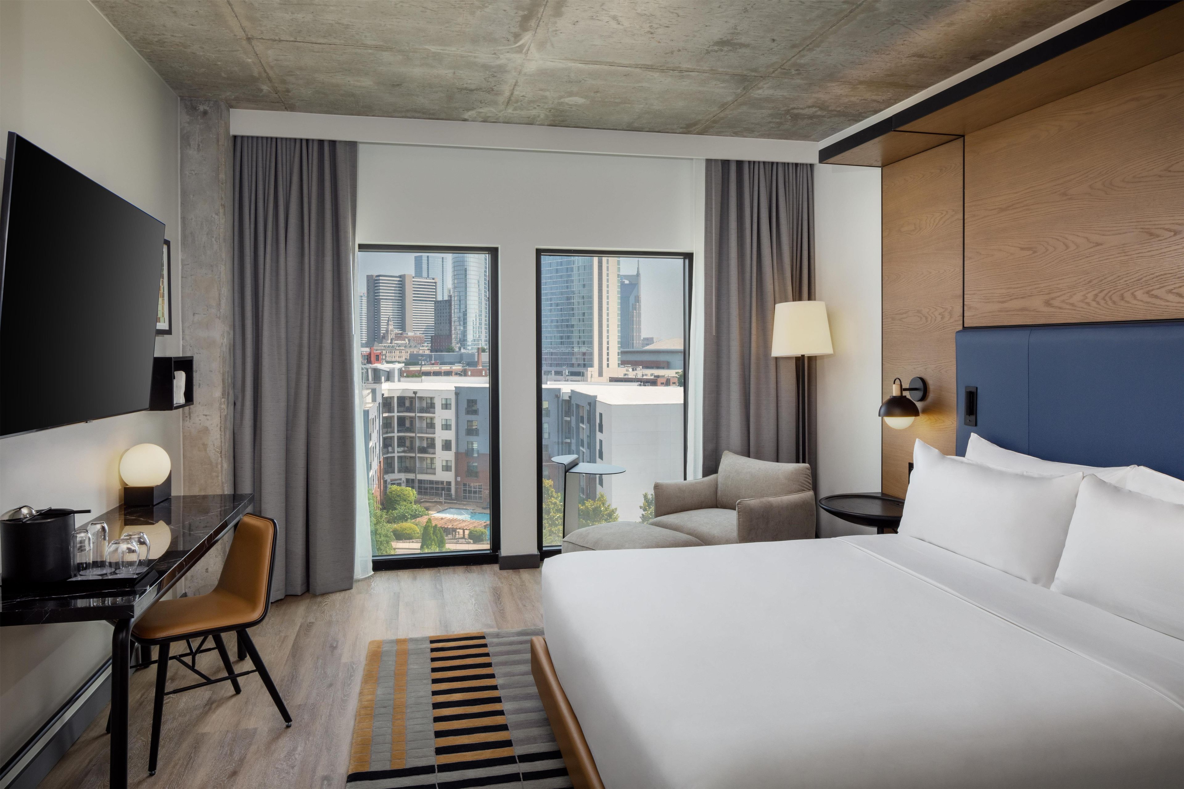 Une chambre d’hôtel avec un grand lit, un bureau avec une chaise, un téléviseur fixé au mur et une vue sur le paysage urbain par les fenêtres.
