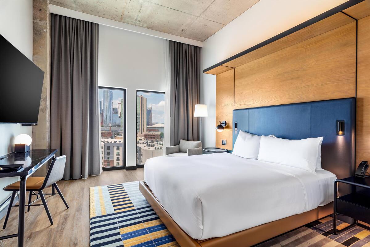 A modern hotel room with a large bed, a desk, and a chair. There is a view of the city through large windows.