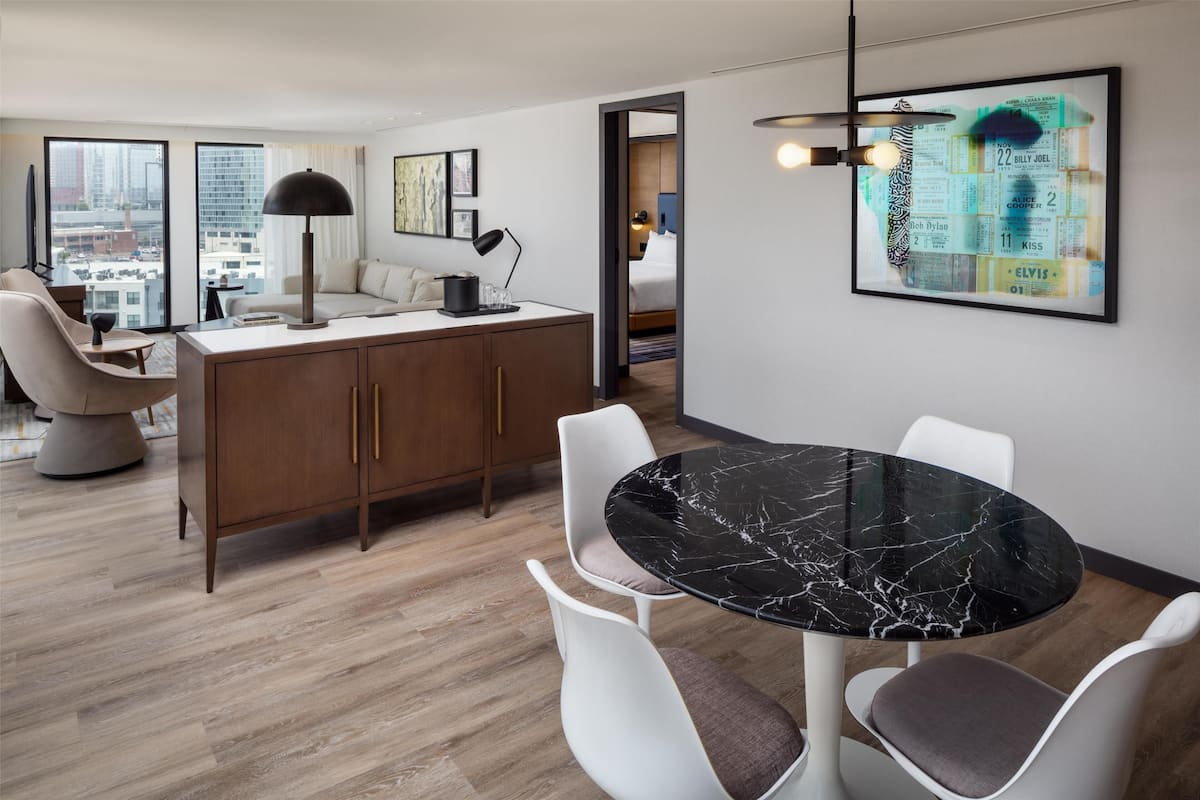 A modern hotel room with a dining area, a sofa, a desk, and a large window offering a city view.
