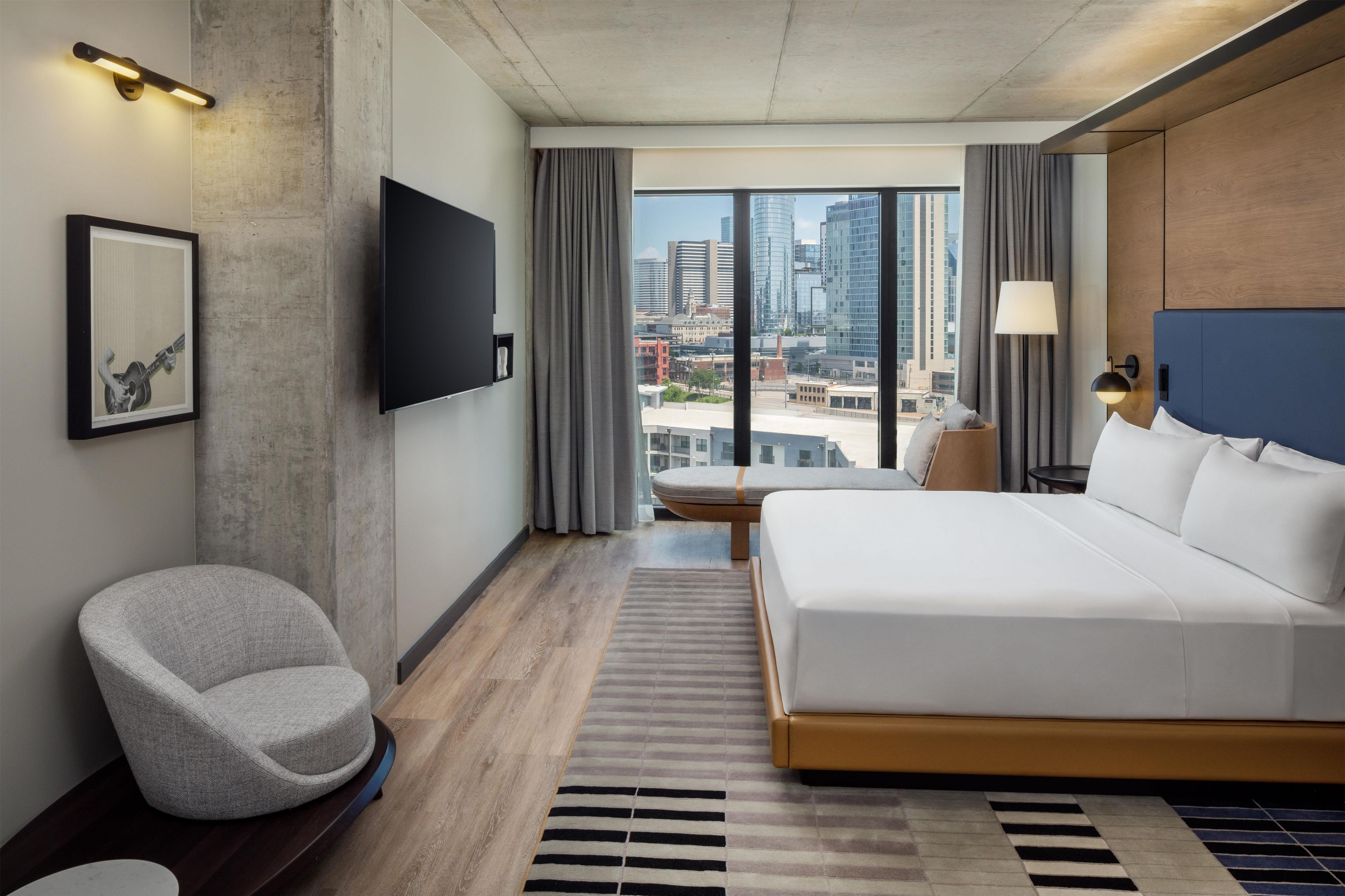 Een moderne hotelkamer met een groot bed, een flatscreen televisie, een grijze fauteuil en uitzicht op de stad.