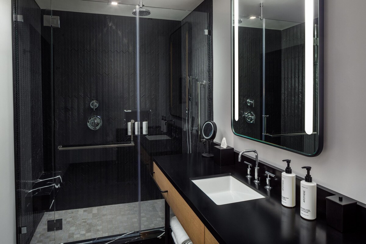 Un baño moderno con ducha envuelta en vidrio, un espejo grande y un mostrador negro con dos lavamanos.