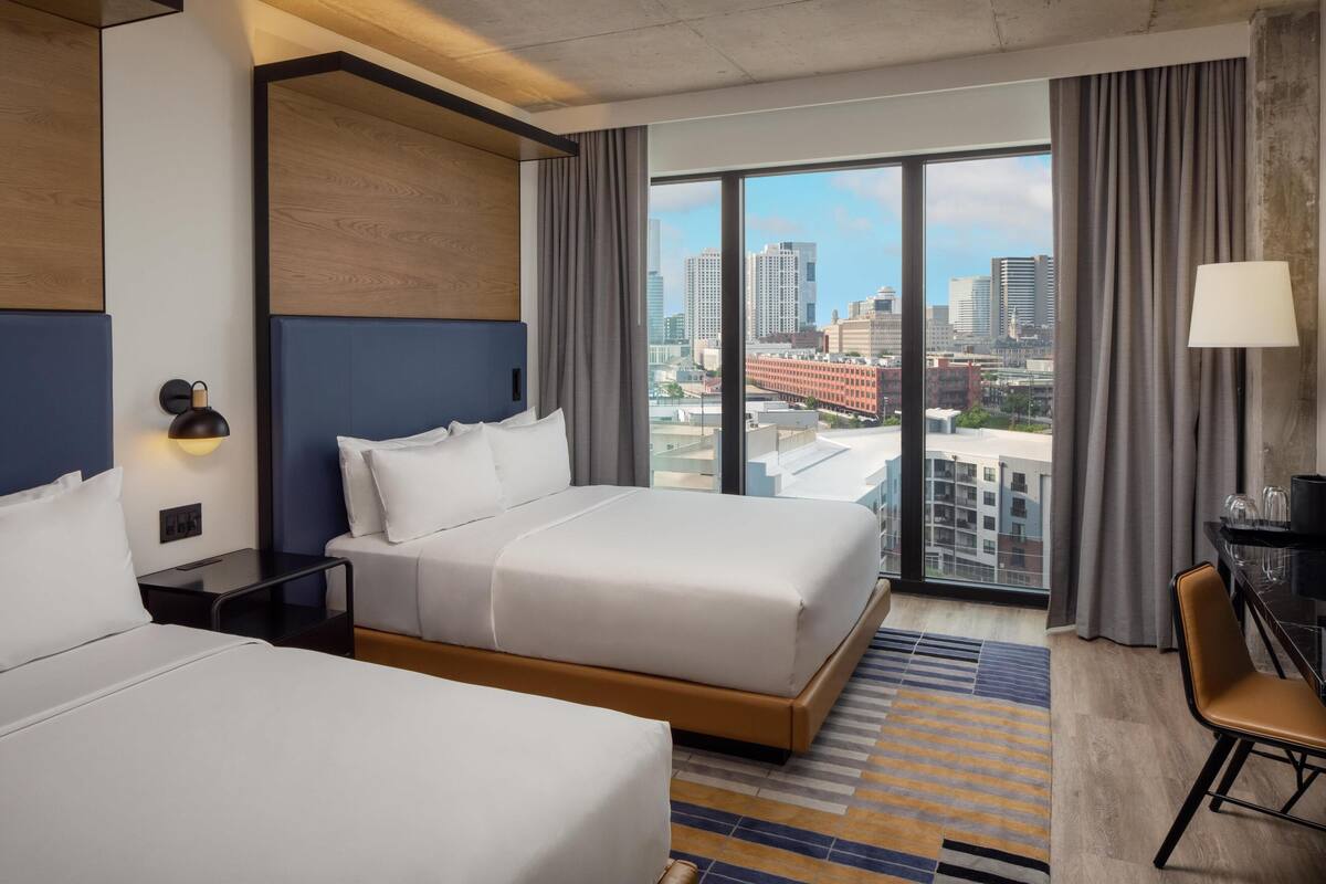 A hotel room with two beds, a large window with a city view, a desk with a lamp, and a chair.
