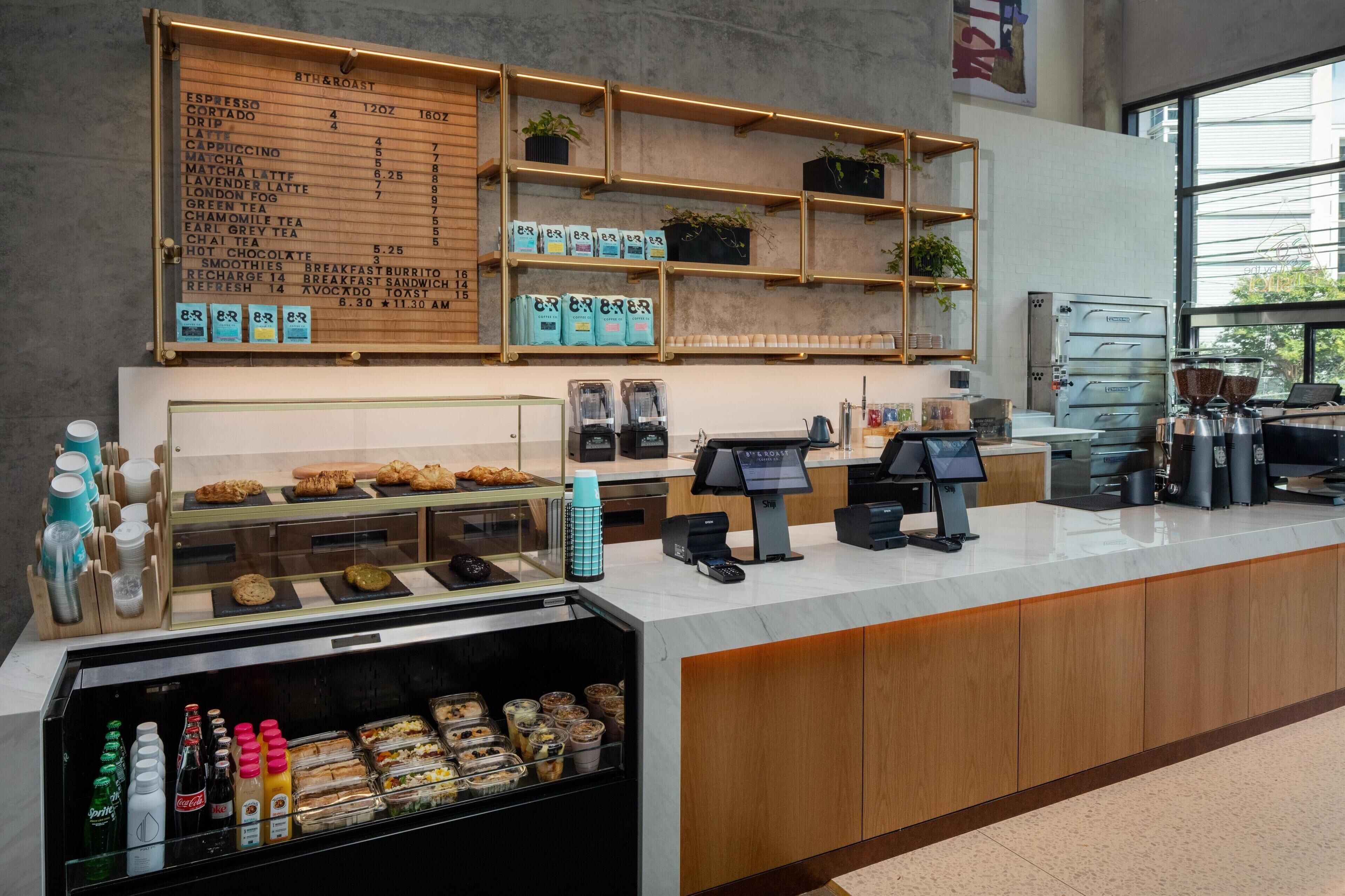 A coffee shop counter with a menu board, beverage dispensers, and a refrigerated display of pastries and sandwiches.