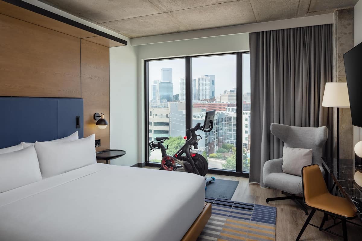 A hotel room with a bed, a chair, a desk, a lamp, and a view of the cityscape through large windows.