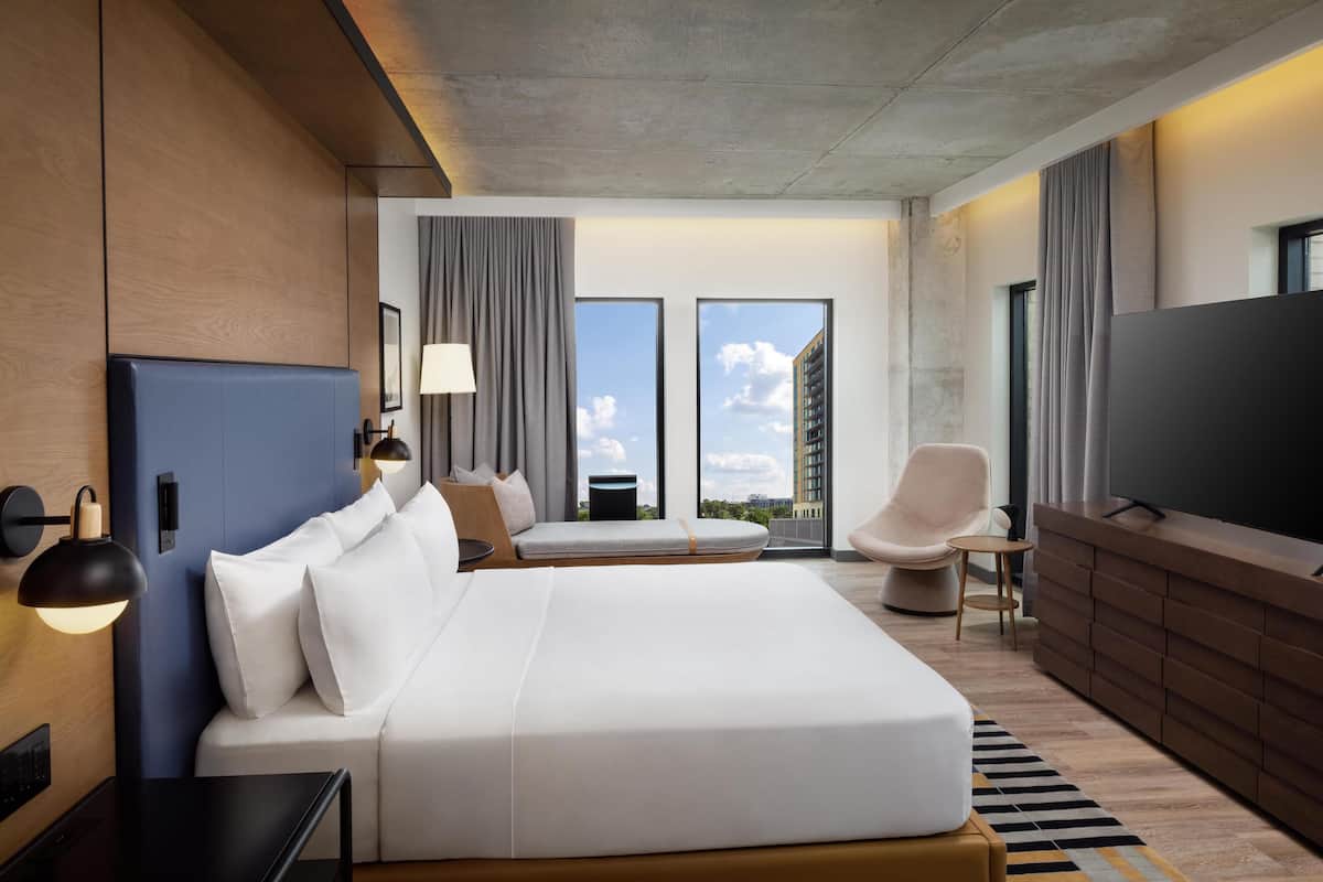 A modern hotel room with a large bed, a TV, a seating area, and a view of the city.