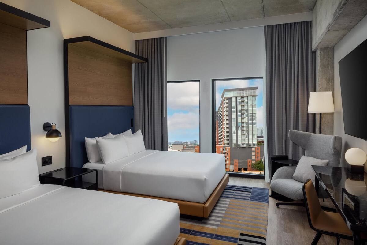 A hotel room with two beds, a desk, a chair, a lamp, and a view of a cityscape.