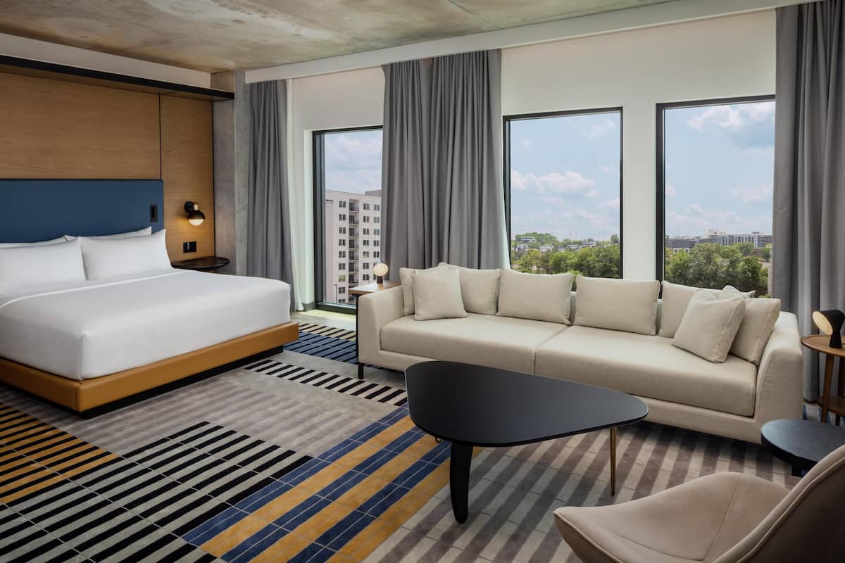 A modern hotel room with a large bed, a sofa, a coffee table, and a view of the city through large windows.