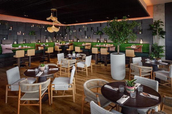 A restaurant with round tables and chairs, a green cushioned bench area, and a wall decorated with plants and shelves.