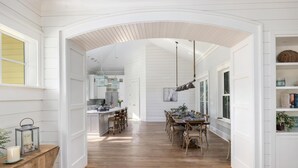A spacious dining area with a long wooden table and chairs, white paneled walls, and a kitchen visible in the background. - 5BR 5BA Luxury Home with Private Pool on IOP! (Isle of Palms)