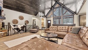 A spacious living room with a large leather sectional sofa, a round coffee table, and a fireplace. There are large windows providing natural light and a view of the outdoors. - Expansive 7 bedroom, 3 car garage, 2 hot tubs, paved roads, wifi & pool access (Lead)