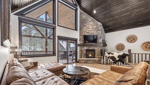 A spacious living area with a stone fireplace, leather sofas, and large windows offering views of the outdoors. - Expansive 7 bedroom, 3 car garage, 2 hot tubs, paved roads, wifi & pool access (Lead)