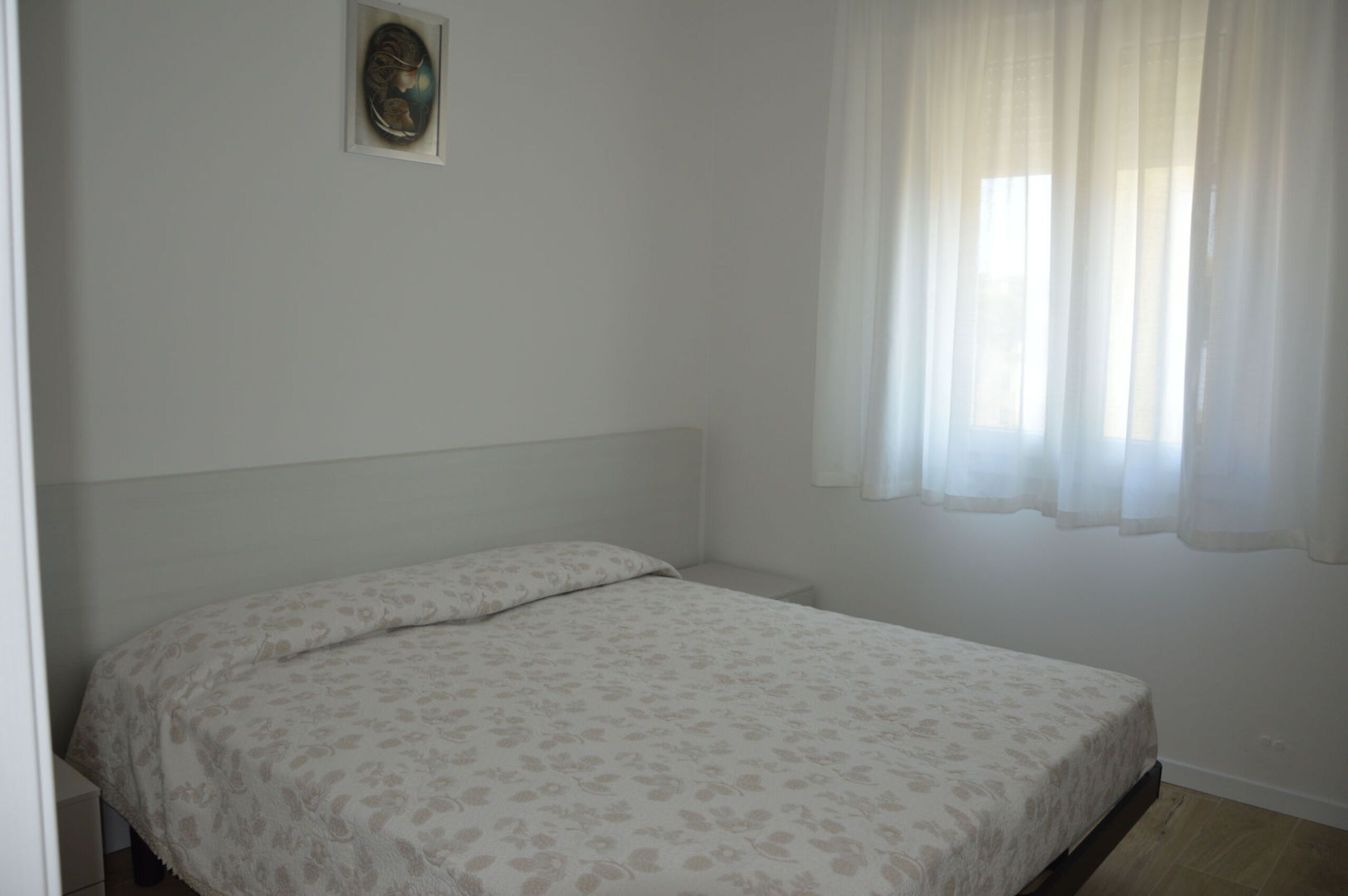 A single bed with a floral-patterned bedspread in a room with white walls and a window with sheer curtains.