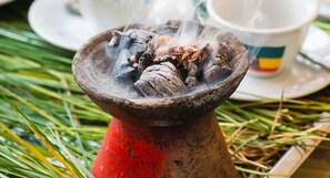 A traditional smoking device with coals and herbs, placed on a red stand, surrounded by green foliage.