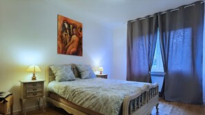 Un dormitorio con cama, dos mesitas de noche con lámparas, un cuadro en la pared y una ventana con cortinas.