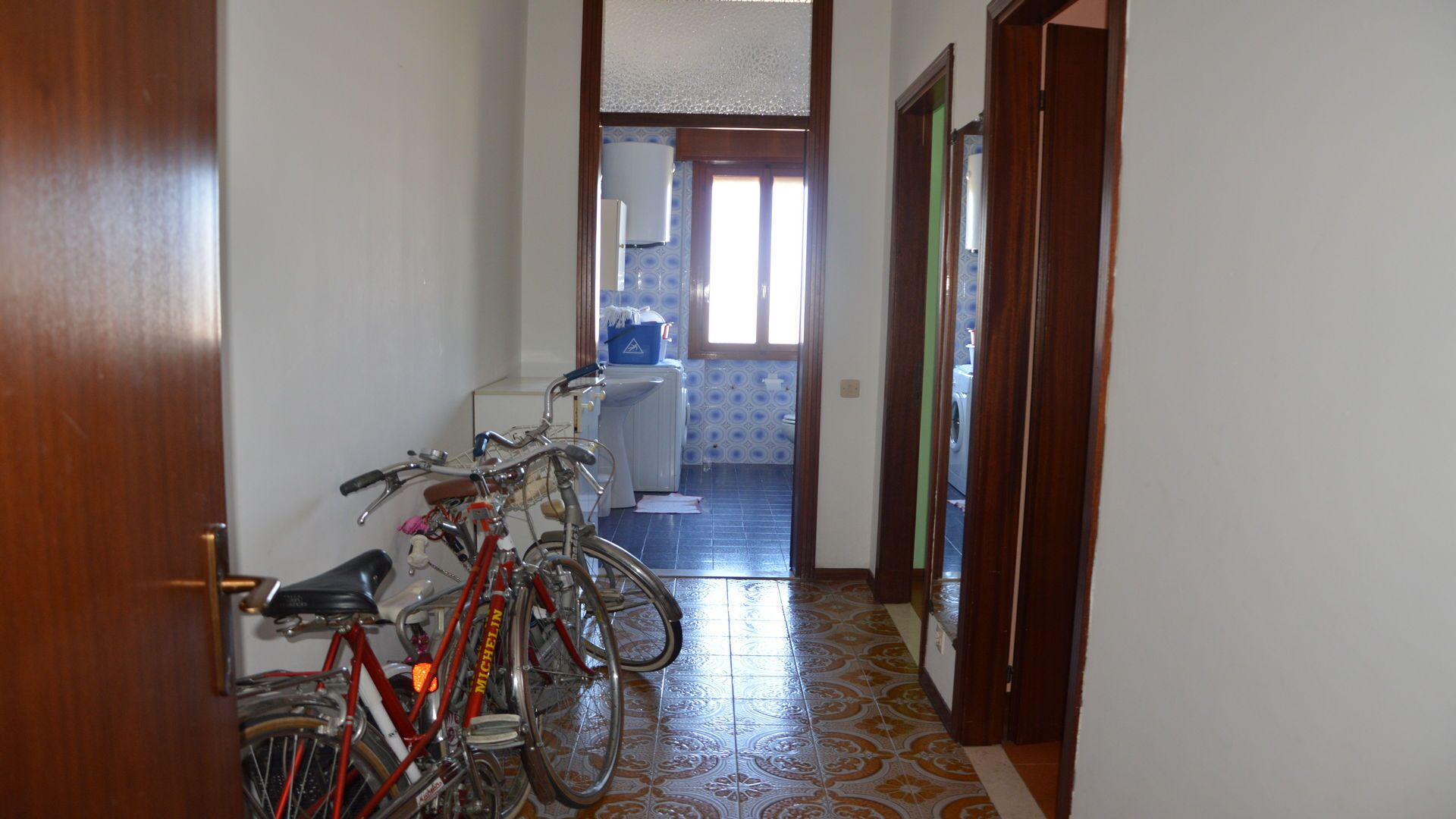 A narrow hallway with a wooden door on the left, leading to a room with a washing machine and a window.
