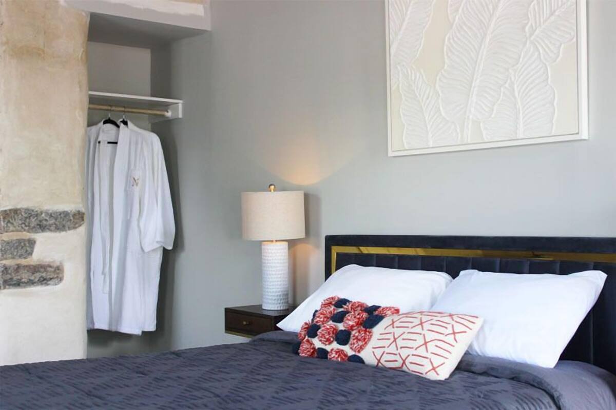 A bedroom with a bed, a nightstand with a lamp, a wardrobe with hanging clothes, and a wall with a leaf pattern.