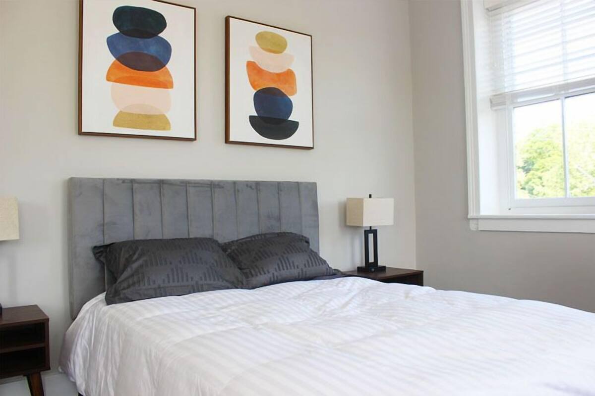 A bedroom with a bed, two nightstands, a window with blinds, and two abstract paintings on the wall.
