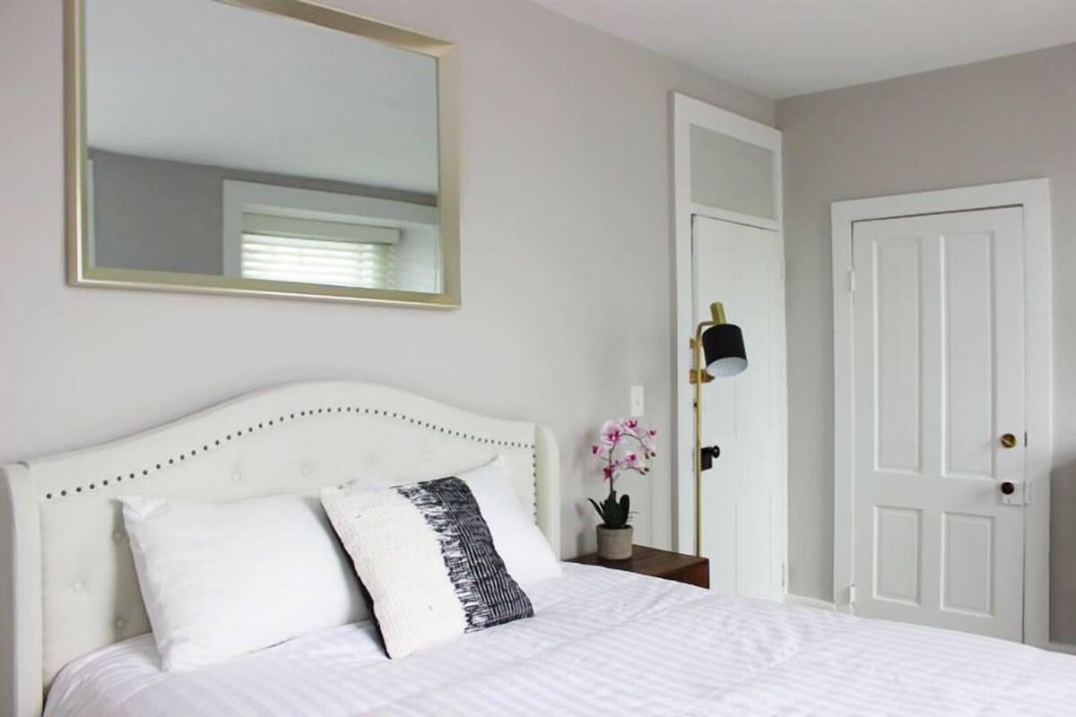 A neatly made bed with white bedding and two pillows, a bedside table with a lamp, a mirror on the wall, and a white door.