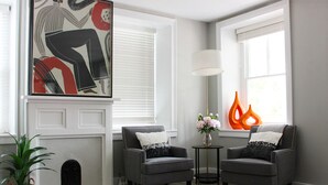 Een moderne woonkamer met twee grijze fauteuils, een witte open haard, een groot abstract schilderij, een staande lamp en een vaas met bloemen.