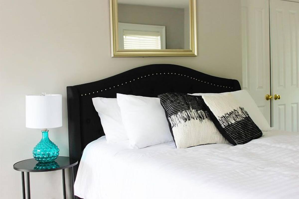 A neatly made bed with white linens and black and white patterned pillows, a teal lamp on a small side table, and a gold-framed mirror above the bed.