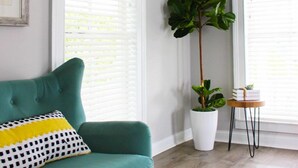 Een smaragdgroene bank met een gekleurd kussen, een hoge potplant, en een klein houten tafeltje met boeken en een plantje naast een raam met witte blinds.