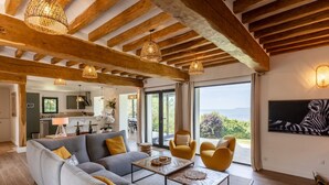 Un salon moderne avec un canapé, des fauteuils et une table basse. On y trouve des luminaires suspendus, un tableau représentant une zèbre et une vue sur la mer par de grandes fenêtres.