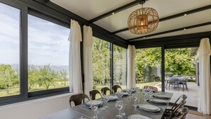 Un coin repas avec une table dressée pour six personnes, de grandes fenêtres offrant une vue sur un vignoble et un plan d’eau.