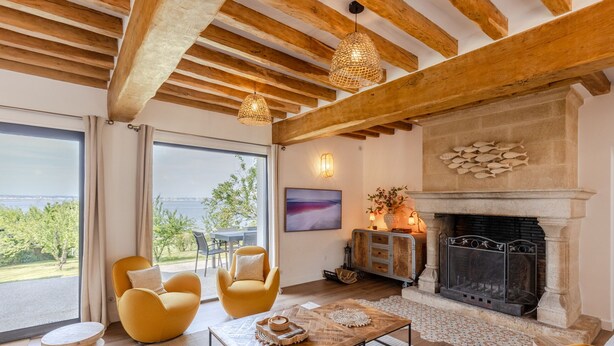 Une pièce avec une cheminée, des poutres apparentes en bois au plafond, une grande fenêtre donnant sur un beau paysage et des fauteuils jaunes.