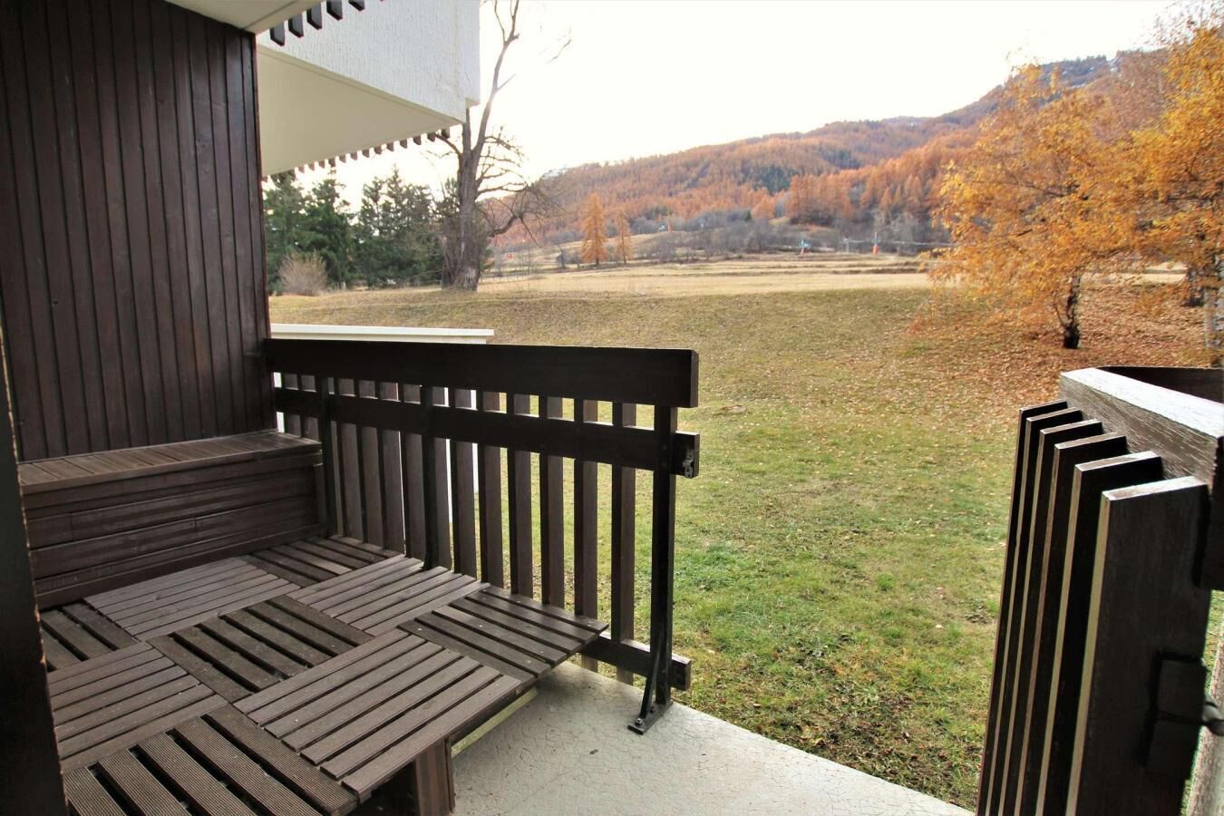 Un banco de madera en un porche con vista a un campo cubierto de hierba y árboles al fondo.