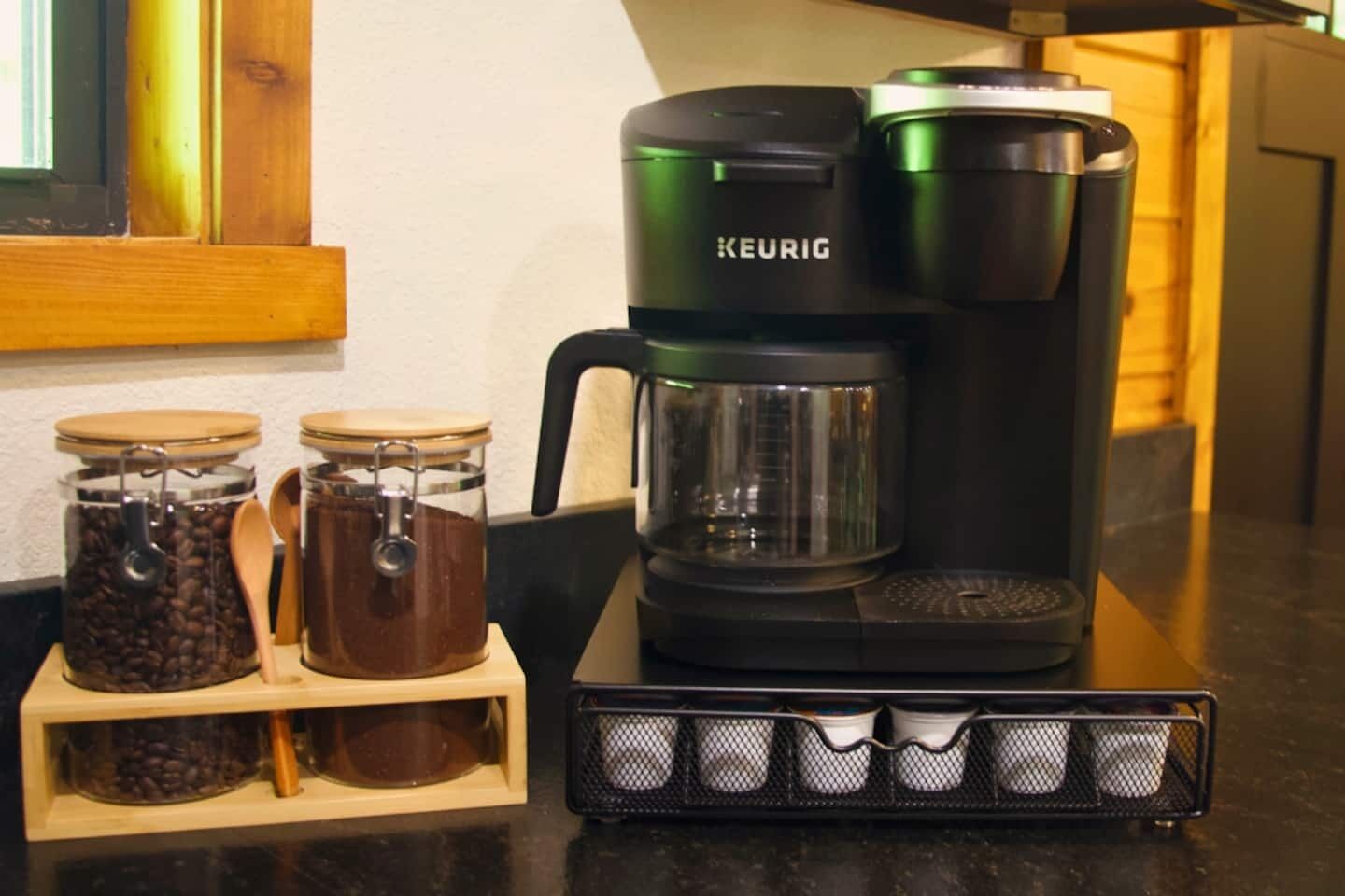 En Keurig kaffemaskine på en køkkenbordplade med to glasbeholdere med kaffebønner og en metalkande med en ske.