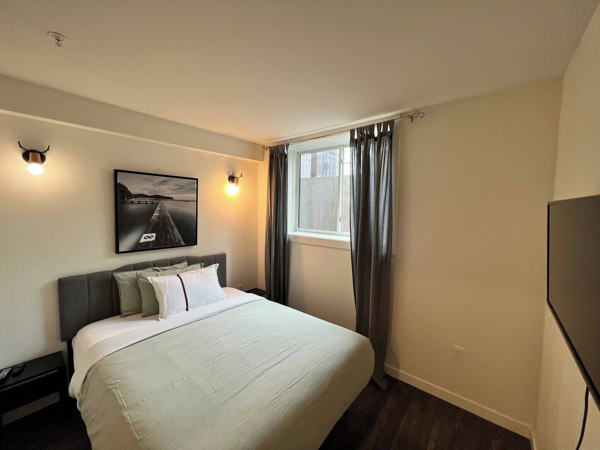 Habitación de hotel con cama, televisión montada en la pared, ventana con cortinas e imagen decorativa en la pared.