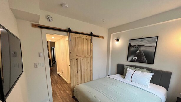 A bedroom with a bed, a wooden door, a framed picture, and a hallway leading to another room. - Dt 1bed 1bath Cozy Suite 8 Min Walk To Arena (Edmonton)
