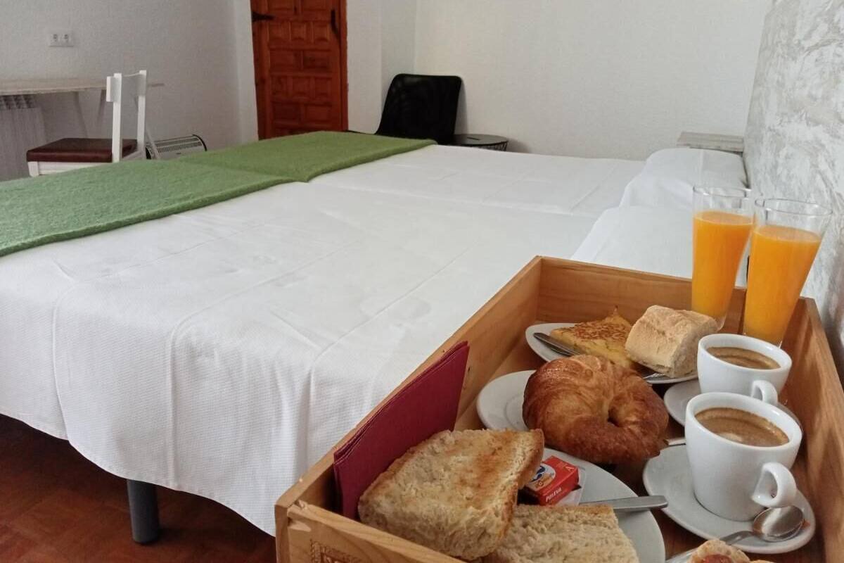 A hotel room with a bed, a wooden side table with breakfast items, and a green throw on the bed.