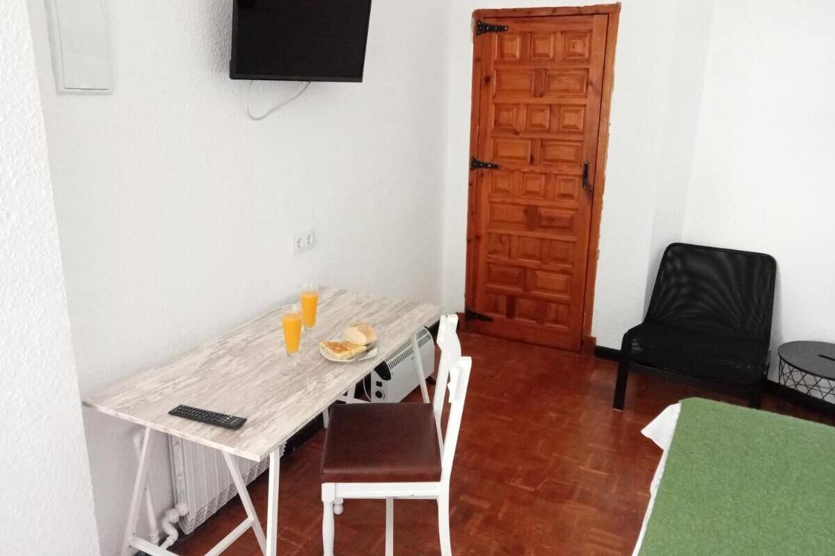 A small room with a wooden door, a TV mounted on the wall, a dining table with chairs, and a small table with a chair.