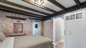 A bedroom with a stone wall, a bed with a beige cover, a door with frosted glass panels, and a small window with a landscape painting.