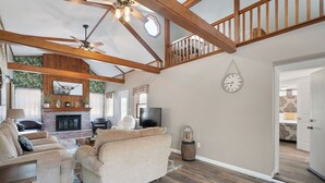 A spacious living room with a high ceiling, wooden beams, a fireplace, a sofa, a television, and a clock on the wall. - In the center of all Historic Main Street has to offer (Grafton)