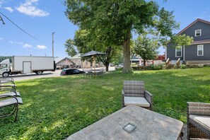 Une cour arrière avec une table à manger, des chaises, un grand arbre et une maison en arrière-plan.