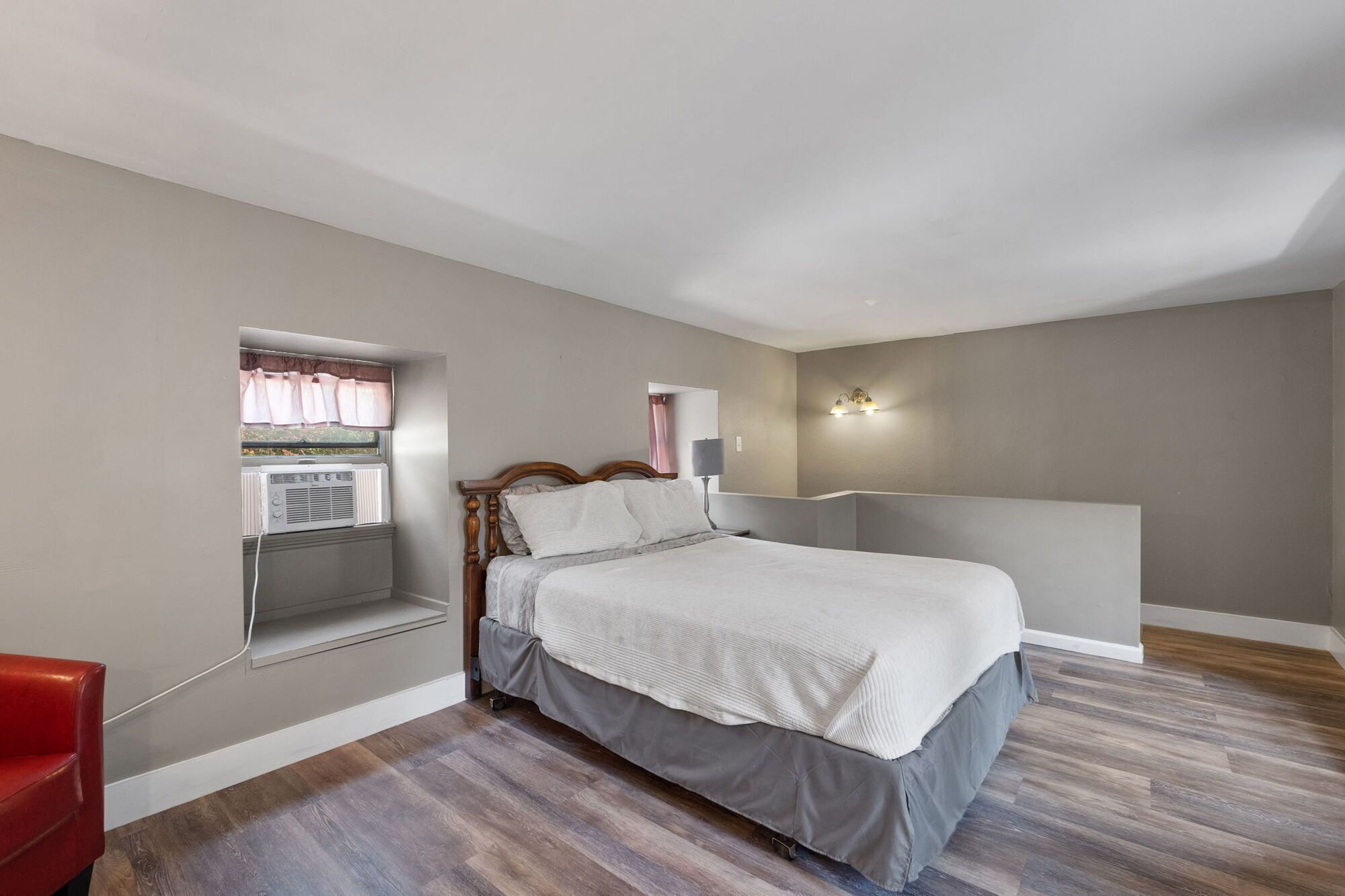 Une chambre à coucher avec un lit, une fenêtre avec des rideaux, un luminaire fixé au mur et une chaise rouge.