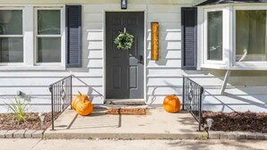 Uma casa com porta, duas janelas com persianas, um tapete na entrada e dois abóboras no alpendre.