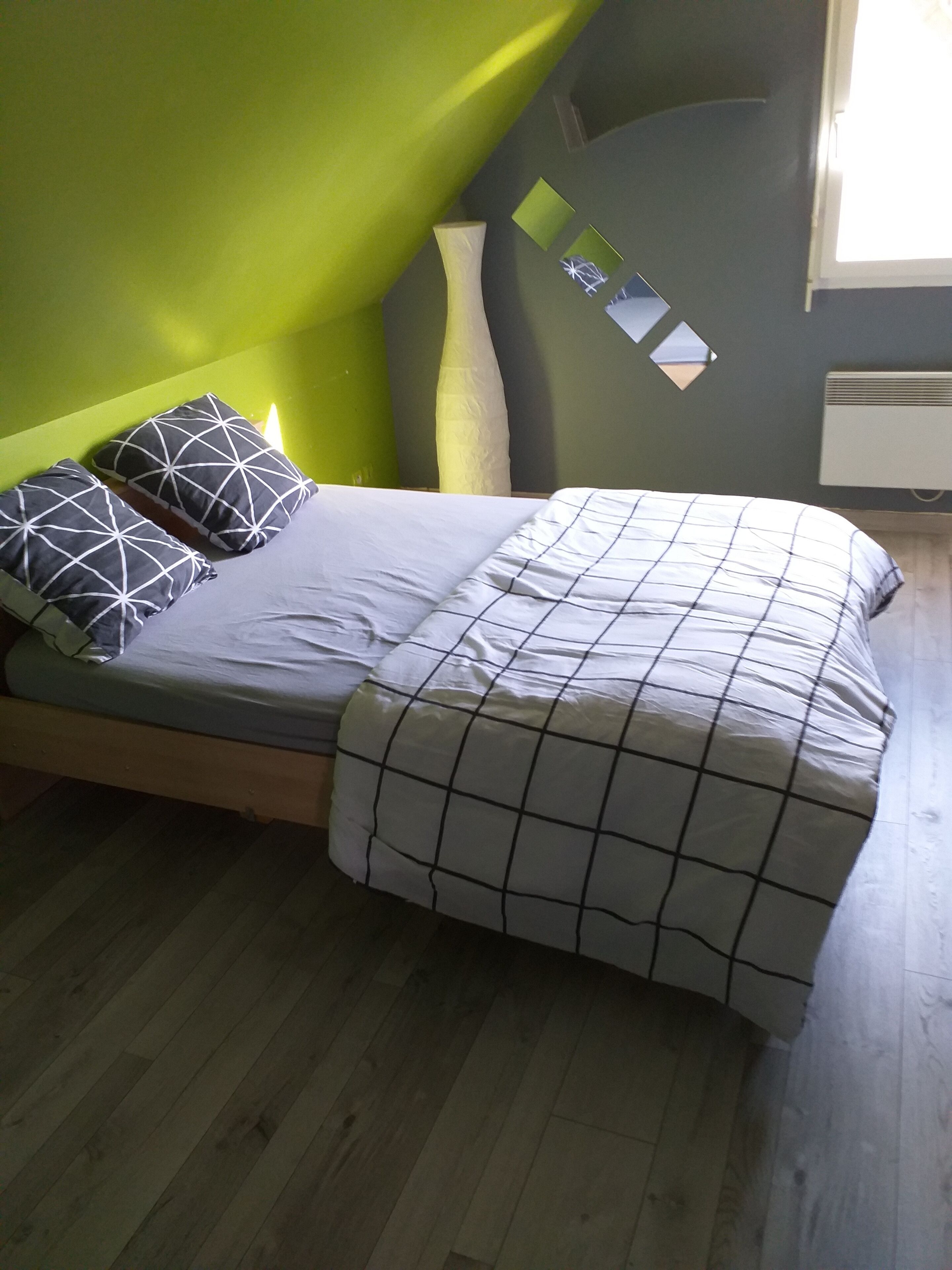 Un lit avec une couvre-lit à carreaux et des coussins aux motifs géométriques, un mur vert et une fenêtre donnant sur un paysage agréable.