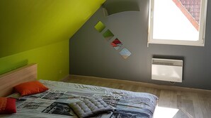Een slaapkamer met een schuine balkenlaag, een bed met een sprei, kussens, een raam met uitzicht op een bakstenen muur en een aan de muur bevestigde airconditioner.