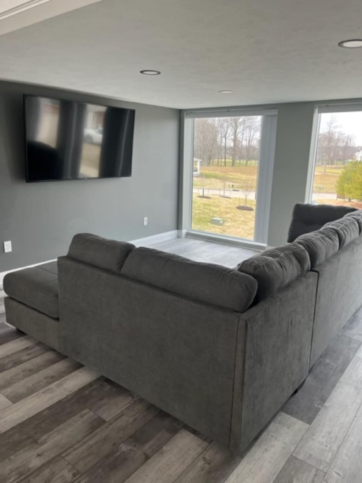 A modern living room with a grey sectional sofa, a flat-screen TV mounted on the wall, and large windows offering a view of the outdoors.