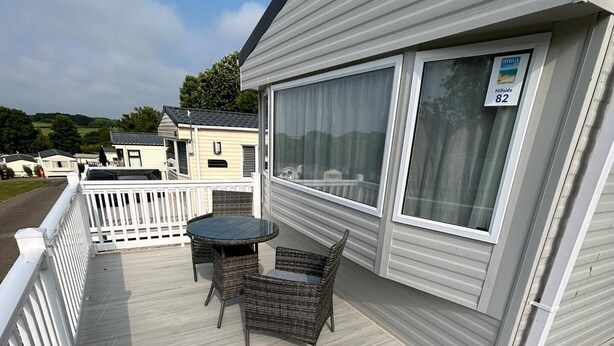 A covered deck with a table and chairs, a white railing, and a window with a sign. - Caravansleeps4-beachfront-garden-patio (Newquay)