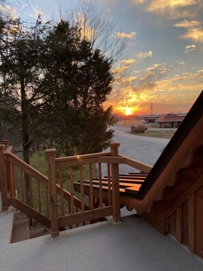 Un escalier en bois mène à une terrasse d’où l’on peut admirer un coucher de soleil sur un paysage rural.