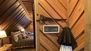 A cozy attic bedroom with a wooden sloped ceiling, a bed with plaid bedding, a 'Welcome' sign, and a coat hanging on the wall. - White Bluff Cabin overlooking Lake Malone  (Lewisburg)